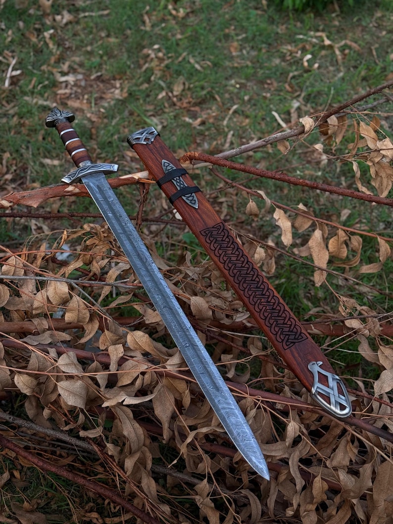 May include: A detailed image of a Viking sword with a silver blade and a wooden handle. The sword is displayed next to its wooden sheath, which features intricate Celtic knotwork and metal accents. The sword and sheath are resting on a bed of twigs and leaves.