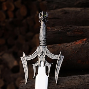 May include: A detailed, silver-colored sword with an ornate handle and a wide, polished blade. The hilt features intricate carvings and a decorative guard. The sword is displayed against a dark, textured wooden background.