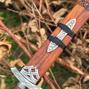 May include: Close-up of a wooden sword sheath with intricate silver metal detailing. The sheath is a rich brown color, with a black cord wrapped around it. The sword's hilt is also visible, featuring similar silver metal accents.