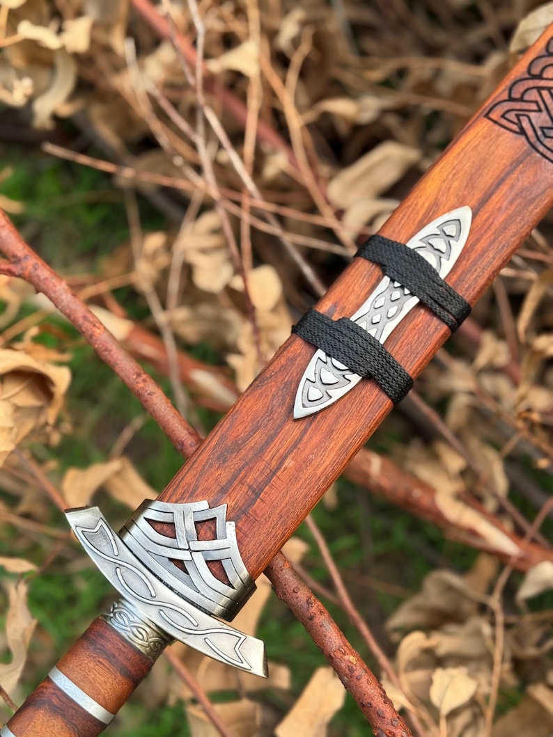 May include: Close-up of a wooden sword sheath with intricate silver metal detailing. The sheath is a rich brown color, with a black cord wrapped around it. The sword's hilt is also visible, featuring similar silver metal accents.