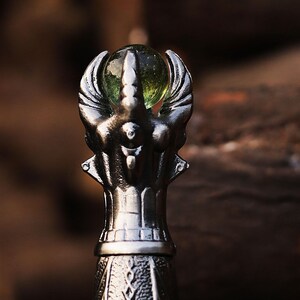 May include: A detailed close-up of a silver-colored bottle stopper. The stopper features an ornate design with a central figure holding a green glass orb. The design includes stylized wings and intricate detailing.