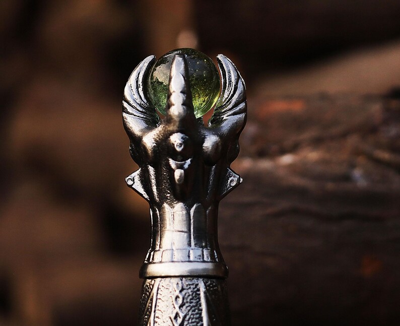May include: A detailed close-up of a silver-colored bottle stopper. The stopper features an ornate design with a central figure holding a green glass orb. The design includes stylized wings and intricate detailing.