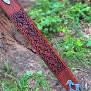 May include: A wooden sword scabbard with a detailed Celtic knot design carved into the reddish-brown wood. The scabbard has a silver-colored metal tip with a bird-like design. The sword's handle is wrapped in black material.