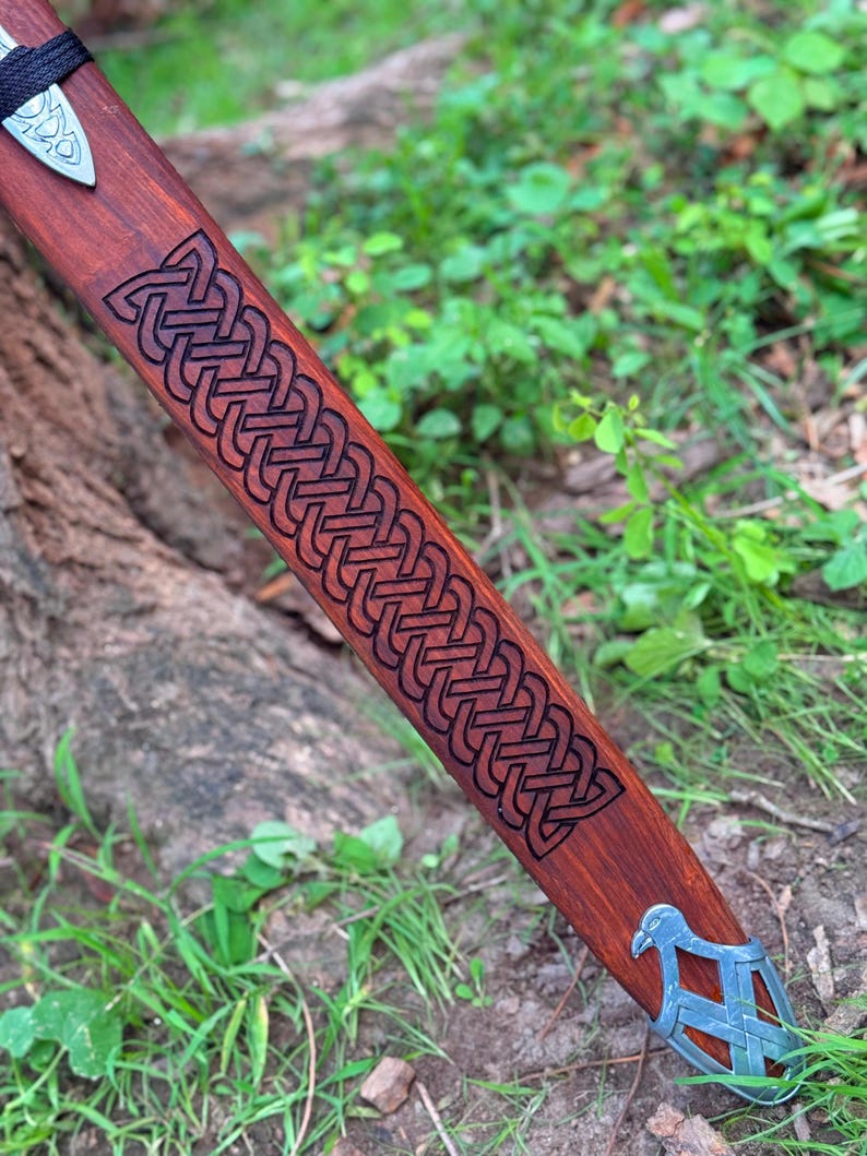 May include: A wooden sword scabbard with a detailed Celtic knot design carved into the reddish-brown wood. The scabbard has a silver-colored metal tip with a bird-like design. The sword's handle is wrapped in black material.
