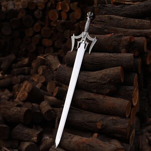 May include: A silver and steel sword with an ornate handle and a long, straight blade. The sword is displayed against a backdrop of stacked, dark brown logs, creating a rustic and dramatic contrast. The handle features intricate detailing.