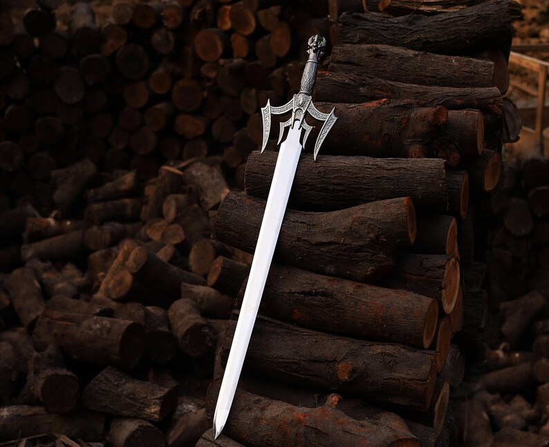 May include: A silver and steel sword with an ornate handle and a long, straight blade. The sword is displayed against a backdrop of stacked, dark brown logs, creating a rustic and dramatic contrast. The handle features intricate detailing.