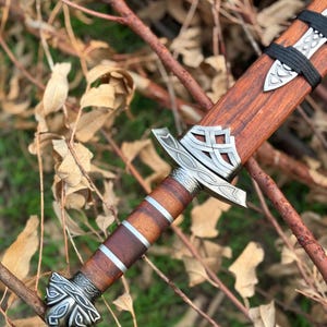 May include: A detailed view of a Viking sword with a wooden handle and sheath. The handle features silver-colored metal accents and a decorative pommel. The blade is not visible. The sword is resting on a bed of twigs and leaves.