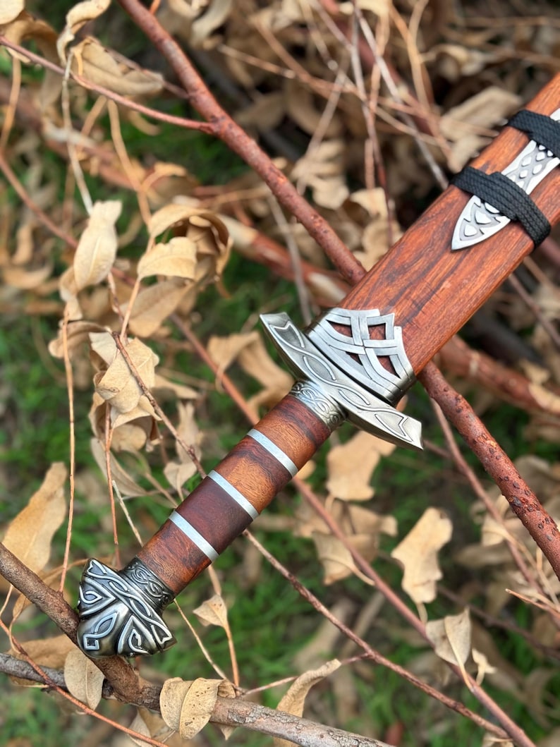 May include: A detailed view of a Viking sword with a wooden handle and sheath. The handle features silver-colored metal accents and a decorative pommel. The blade is not visible. The sword is resting on a bed of twigs and leaves.