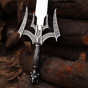 May include: A detailed close-up of a silver sword with an ornate handle and blade. The sword features intricate designs on the hilt and guard, with a polished, reflective blade. The sword is set against a backdrop of dark brown wood.