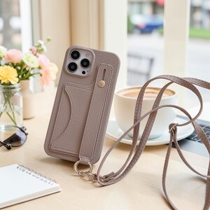 May include: A taupe-coloured textured phone case with a card slot and a vertical strap featuring a gold-tone metal stud. The case includes a matching long crossbody strap attached to a circular metal ring at the bottom, designed for mobile devices.