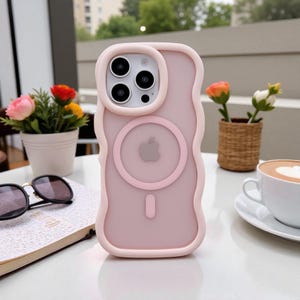 Puede incluir: Funda de teléfono rosa claro con un diseño de borde ondulado. La funda tiene un cargador MagSafe circular y un recorte para la cámara. El logotipo de Apple es visible en la parte posterior. La funda está sobre una mesa blanca, con flores y una taza de café.