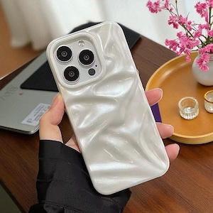 May include: A pearlescent white phone case with a wavy, textured design. The case has a camera cutout with three lenses and a flash. The phone case is held in a hand.