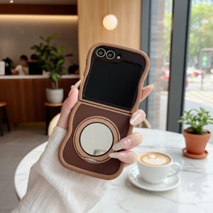 May include: A brown and tan phone case with a mirror and camera cutouts. The case is designed for a folding phone, with a rounded shape and a contrasting border. The phone case is open, revealing the phone's screen.