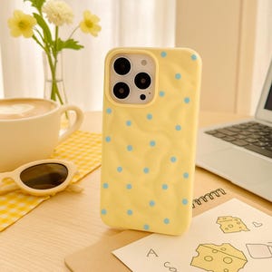 May include: A yellow iPhone case with a pattern of small blue polka dots. The case has a marble-like texture and a camera cutout. The case is on a wooden surface next to a cup of coffee, sunglasses, and a notepad.