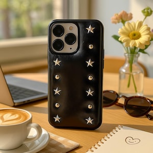 Könnte beinhalten: Schwarze Handyhülle mit silbernen Sternen und Nieten. Die Hülle liegt auf einem Holztisch mit einem Laptop, einer Tasse Kaffee, einer Sonnenbrille und einer kleinen Vase mit Blumen im Hintergrund.