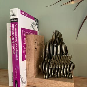 May include: A silver metal sculpture of a person reading, set on a wooden bookend. The bookend supports two books, one titled "Bitcoin & Blockchain". The sculpture is silver and the books are white and purple. The bookend is made of wood.