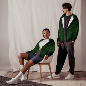 May include: Two people wearing matching green and black zip-up hoodies with a digital rain pattern. The person on the left is seated, wearing a white top, blue shorts, and white sneakers. The person on the right is standing, wearing a gray shirt and dark gray sweatpants.