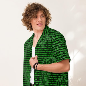 Puede incluir: Una camisa de manga corta con botones frontales que presenta un fondo negro con un patr&oacute;n repetitivo de d&iacute;gitos de c&oacute;digo binario en color verde brillante. La camisa tiene un cuello cl&aacute;sico y se lleva sobre una camiseta blanca sin mangas.