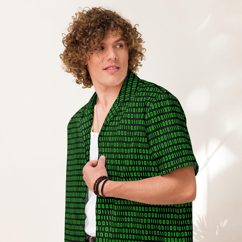 Puede incluir: Una camisa de manga corta con botones frontales que presenta un fondo negro con un patr&oacute;n repetitivo de d&iacute;gitos de c&oacute;digo binario en color verde brillante. La camisa tiene un cuello cl&aacute;sico y se lleva sobre una camiseta blanca sin mangas.