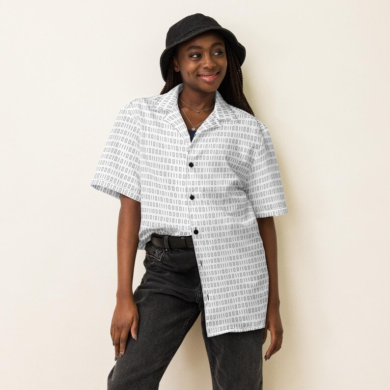 Op de afbeelding: Een overhemd met korte mouwen en knoopsluiting, voorzien van een herhalend binair codepatroon in donkergrijs op een witte achtergrond, gecombineerd met een zwarte bucket hat en een donkergrijze spijkerbroek. Het overhemd heeft een klassieke kraag.
