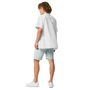 Op de afbeelding: Een overhemd met korte mouwen en knoopsluiting met een herhalend patroon van verticale donkergrijze binaire codecijfers op een witte achtergrond, gecombineerd met een effen lichtgrijze korte broek en witte sneakers, gezien van achteren.