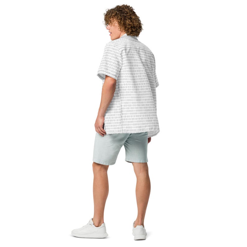 Op de afbeelding: Een overhemd met korte mouwen en knoopsluiting met een herhalend patroon van verticale donkergrijze binaire codecijfers op een witte achtergrond, gecombineerd met een effen lichtgrijze korte broek en witte sneakers, gezien van achteren.