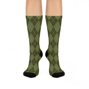 May include: A pair of mid-calf socks featuring an olive green and dark forest green argyle diamond pattern with thin white intersecting lines. The socks have solid black toe and heel sections, providing a distinct contrast to the patterned body.