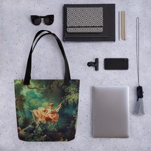 Può includere: Borsa tote nera con un dipinto di una donna su un'altalena in una foresta. L'opera d'arte utilizza tonalità rosa, verdi e blu. La borsa ha manici neri ed è mostrata con occhiali da sole, un taccuino e un computer portatile.