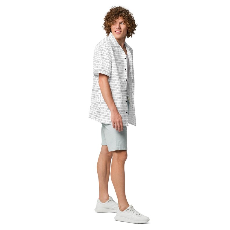 Op de afbeelding: Een overhemd met korte mouwen en knoopsluiting, voorzien van een herhalend patroon van binaire code in het zwart op een witte achtergrond, gecombineerd met een lichtgrijze korte broek en witte sportschoenen. Het overhemd heeft een klassieke kraag.