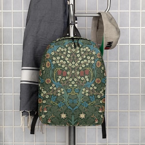 May include: A backpack featuring a dark green background with a dense, symmetrical botanical pattern of white, red, and blue flowers, green leaves, and brown stems. The bag has black straps and zippers, and is displayed hanging on a metal rack.
