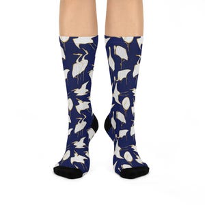 May include: A pair of crew-length socks featuring a dark navy blue background with a repeating pattern of white heron birds outlined in gold. The socks have solid black toe and heel sections, providing a distinct contrast to the patterned fabric.