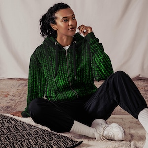 Puede incluir: Una sudadera con cremallera negra con un patrón de lluvia digital verde. La sudadera tiene una cremallera negra y se ve una camiseta interior blanca. La persona lleva pantalones negros y zapatillas blancas. El tema general es la tecnología y la ciencia ficción.