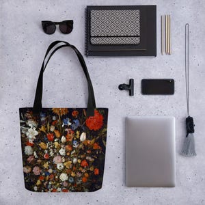 May include: Black tote bag featuring a vibrant floral still life painting. The artwork showcases a dense arrangement of colorful flowers, including red, white, and yellow blooms, set against a dark background. The bag has black handles and is displayed with other accessories.