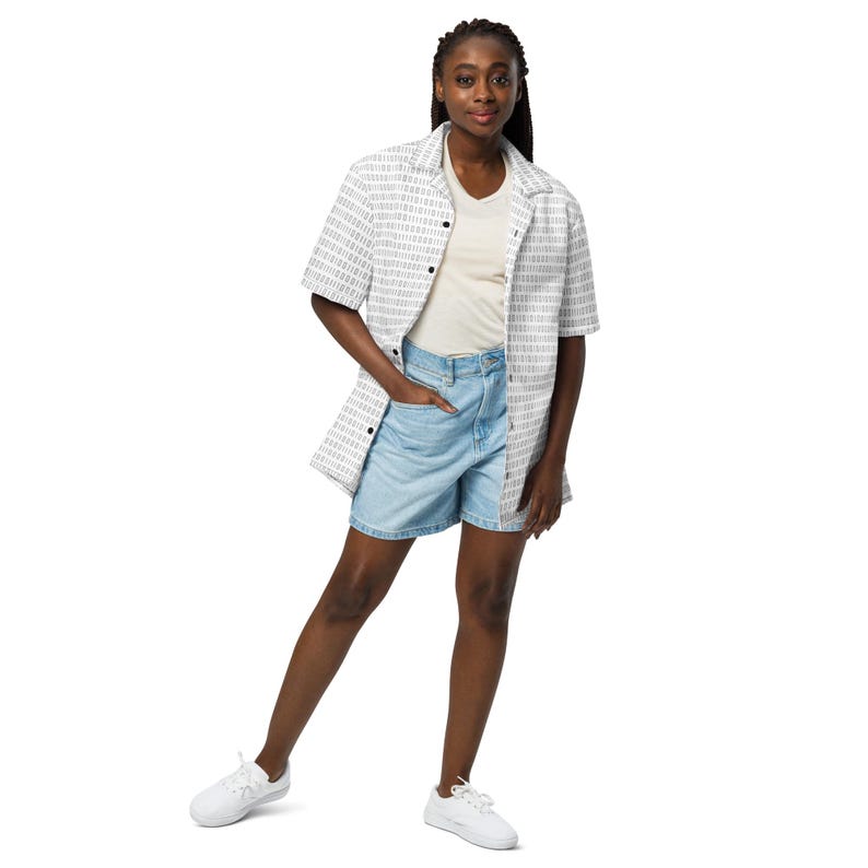Op de afbeelding: Een overhemd met korte mouwen en knoopsluiting met een wit en grijs binair codepatroon, gedragen over een effen cr&egrave;mekleurig T-shirt met V-hals, lichtblauwe spijkershorts en witte sneakers. Het geheel is casual en heeft een klassieke kraag.