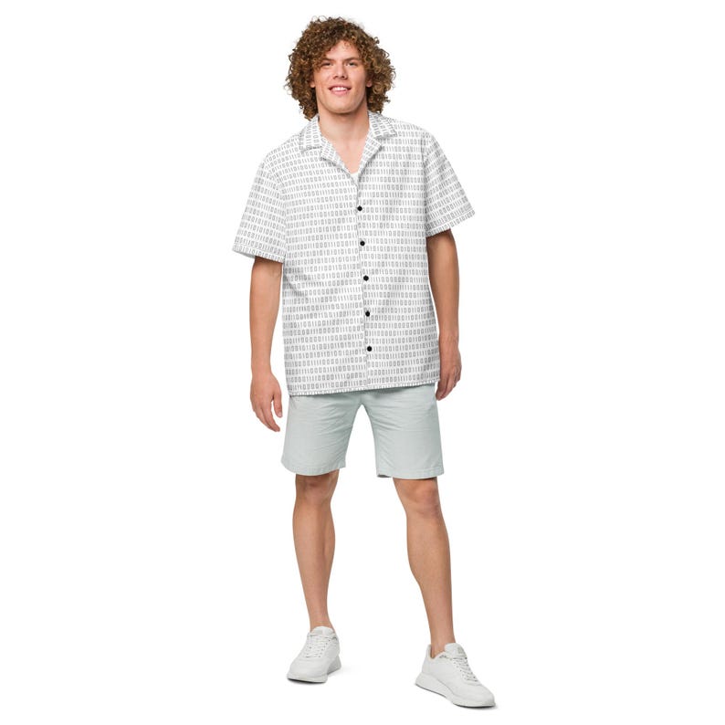 Op de afbeelding: Een overhemd met korte mouwen en knoopsluiting, voorzien van een herhalend binair codepatroon van donkergrijze nullen en enen op een witte achtergrond, gecombineerd met een lichtgrijze korte broek en witte sportschoenen. Het overhemd heeft een klassieke kraag.