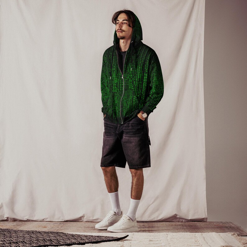 May include: A green and black zip-up hoodie with a matrix-style pattern. The hoodie has a hood and a zipper down the front. The person is wearing black denim shorts, white socks, and white sneakers. A silver watch is on the person's wrist.
