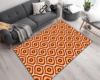 May include: A rectangular area rug with a retro pattern. The rug features a repeating design of stylized, rounded shapes in shades of orange, brown, and cream. The rug is placed on a light-colored wooden floor, near a gray sofa.