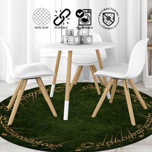 May include: A round, dark green rug with gold lettering and a grass-like texture, placed under a white table and chairs. The table and chairs have light wood legs and white seats. The rug has the words "The Lord of the Rings" in gold lettering.