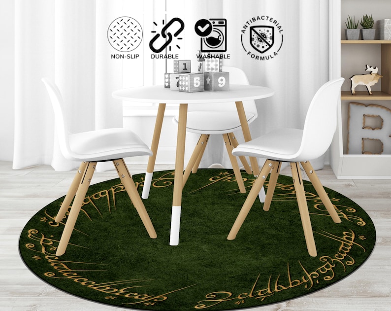 May include: A round, dark green rug with gold lettering and a grass-like texture, placed under a white table and chairs. The table and chairs have light wood legs and white seats. The rug has the words "The Lord of the Rings" in gold lettering.
