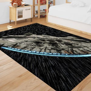 Puede incluir: Una alfombra rectangular con una ilustración detallada de una nave espacial sobre un fondo negro con rayas blancas. La nave espacial es principalmente gris con detalles azules.