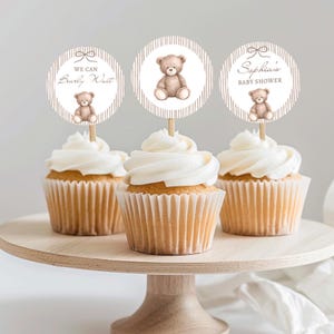 Op de afbeelding: Drie cupcakes met witte glazuur en teddybeer toppers op een houten taartstandaard. De toppers hebben de tekst "We Can Bearly Wait" en "Sophia's Baby Shower". De toppers tonen een bruine teddybeer illustratie.