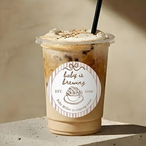 Può includere: Caffè freddo in un bicchiere di plastica trasparente con una cannuccia nera. La bevanda ha un aspetto cremoso e stratificato, guarnito con panna montata e una spolverata di cannella. Un adesivo rotondo sulla tazza recita "baby is brewing".