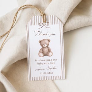 Op de afbeelding: Een cadeau-label voor een babyshower met een aquarel illustratie van een teddybeer. Het label heeft een gestreepte rand en een strik bovenaan. Tekst bevat "Bedankt", "voor het verwennen van onze baby met liefde", een naam en een datum.