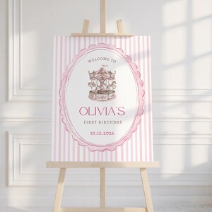 May include: A pink and white striped welcome sign on a wooden easel. The sign features an oval frame with a carousel illustration and the text "WELCOME TO OLIVIA'S FIRST BIRTHDAY 10.12.2026".