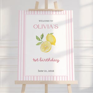 Könnte beinhalten: Ein Willkommensschild auf einer hölzernen Staffelei mit rosa und weißen Streifen. Das Schild zeigt "Welcome to Olivia's" in rosa Schrift, mit einer Aquarellillustration von Zitronen. Der Text "1. Geburtstag" und das Datum "21. Juni 2026" sind ebenfalls angezeigt.