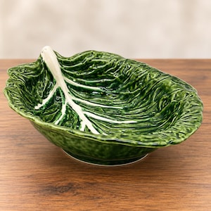 May include: A glossy ceramic bowl shaped like a large, ruffled green cabbage leaf. The bowl features a prominent, raised white central vein that extends from the edge toward the center, with deep, textured green folds mimicking natural foliage.