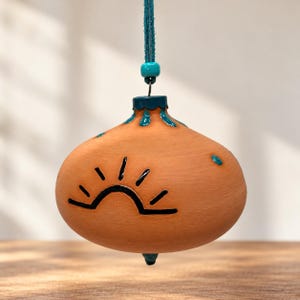 May include: A terracotta-colored ceramic ornament with a rounded shape, featuring a black hand-painted sun graphic. The top and bottom are accented with glossy teal glaze, and it hangs from a teal cord with a matching bead.