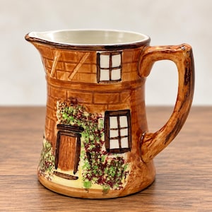 May include: A ceramic pitcher shaped like a cottage with a brown brick pattern, a handle, and a spout. The exterior features painted details of a door, windows with white panes, and green and purple climbing vines against the brown brick walls.