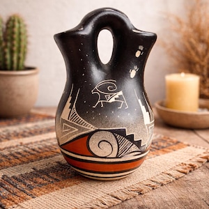 May include: A black ceramic vase with a handle and a decorative design. The vase features a white goat illustration, geometric patterns, and red and black bands. The vase is on a woven mat.
