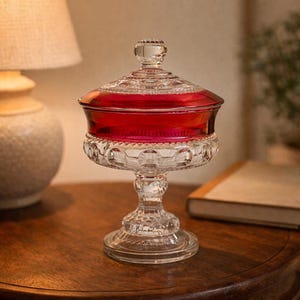 May include: A clear glass candy dish with a ruby red interior and a decorative lid. The dish sits on a pedestal base and is displayed on a wooden table. The dish is likely used for decorative purposes.